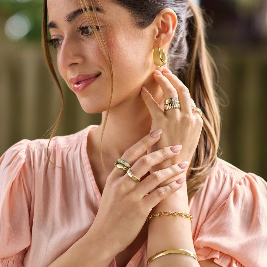 Woman wearing gold jewelry including earrings, rings, and a bracelet with a blurred green background.